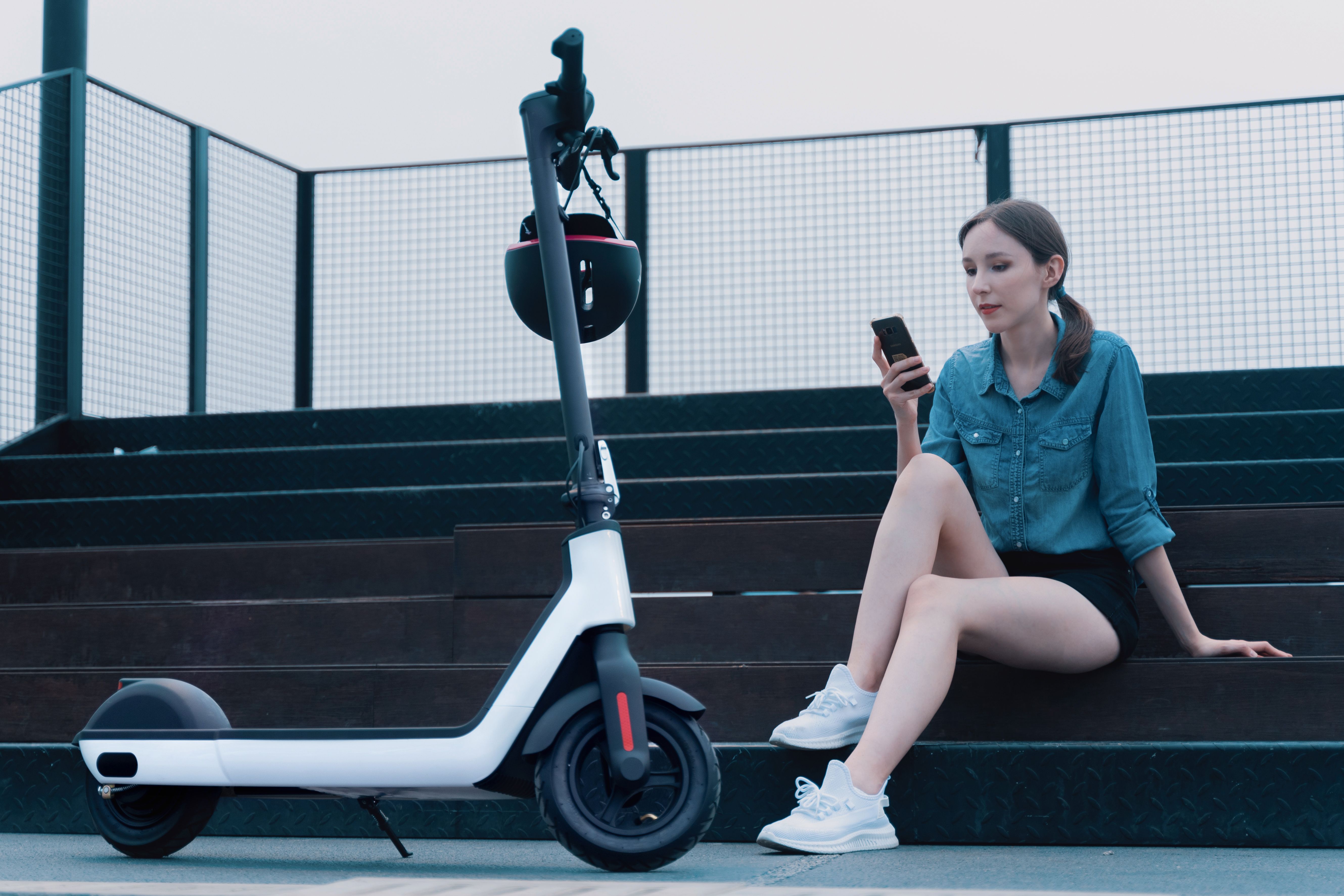Woman sitting beside an electric scooter while researching it on her smartphone